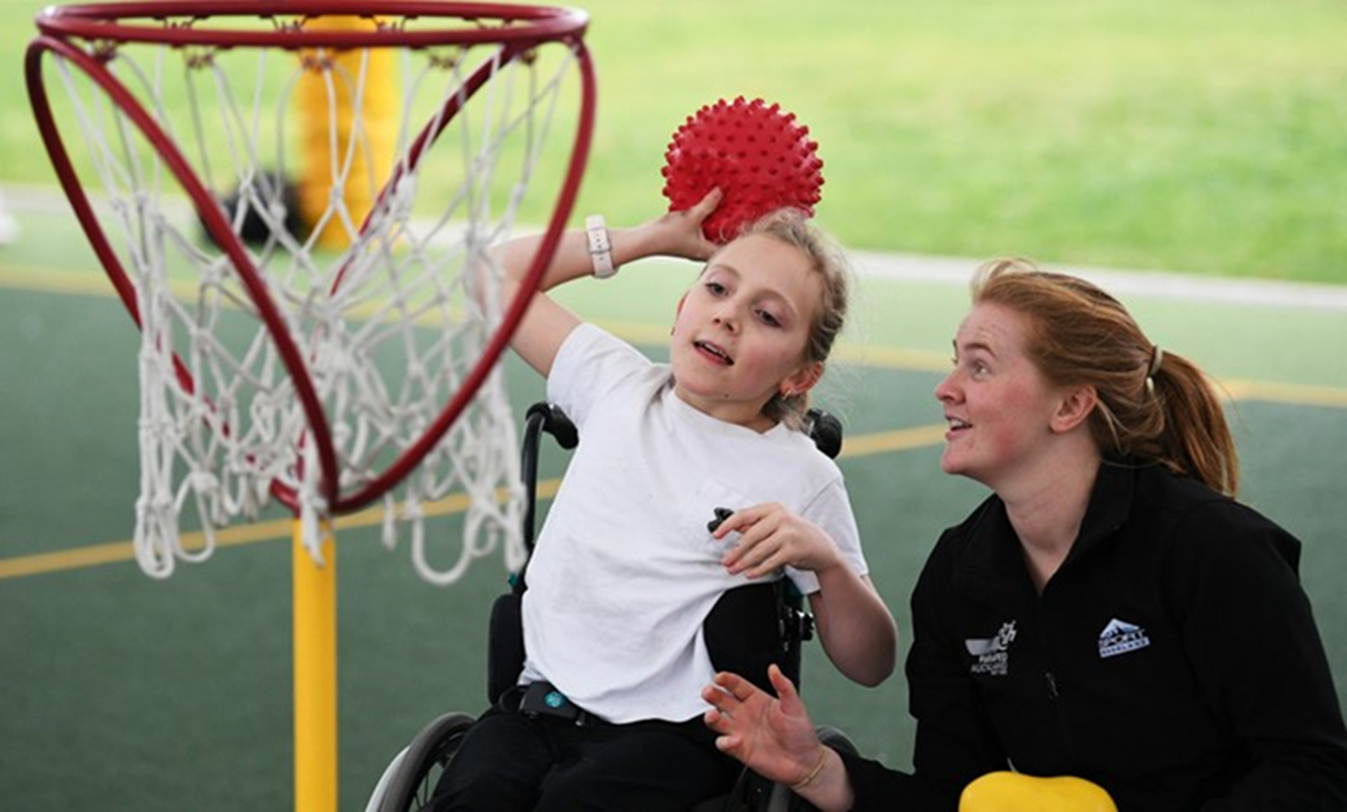 Female coach overlooking young girl in wheelchair throwing a ball into a hoop